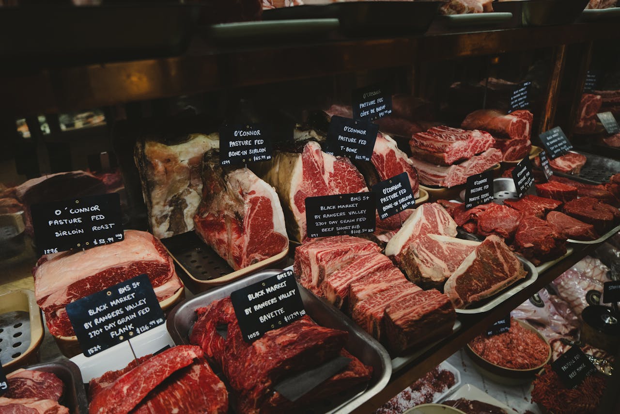 A range of raw meat cuts on display in a butcher shop, featuring labels for each type.